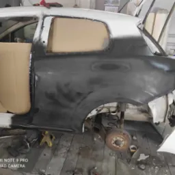 Przygotowanie karoserii samochodu do lakierowania: szpachlowana i oszlifowana powierzchnia boczna, zakryta kartonem szyba, widoczne elementy zawieszenia, podnośnik pod progiem.