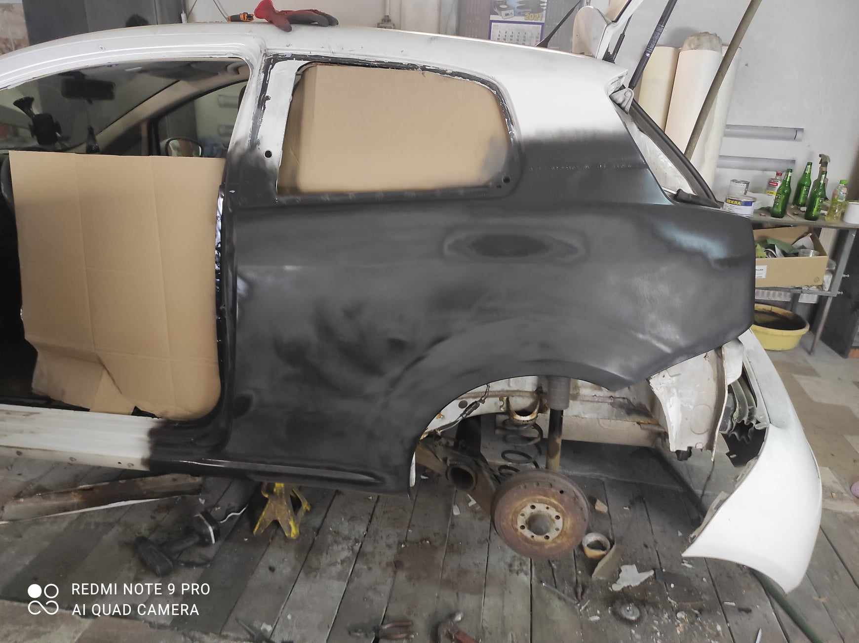 Przygotowanie karoserii samochodu do lakierowania: szpachlowana i oszlifowana powierzchnia boczna, zakryta kartonem szyba, widoczne elementy zawieszenia, podnośnik pod progiem.