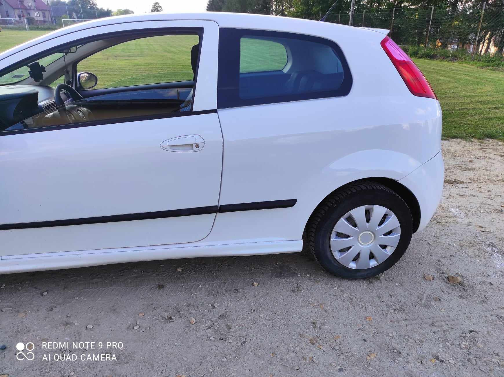 Biały samochód osobowy, widok z boku, lekko zakurzone nadwozie, kołpak na przednim kole, tło zielone