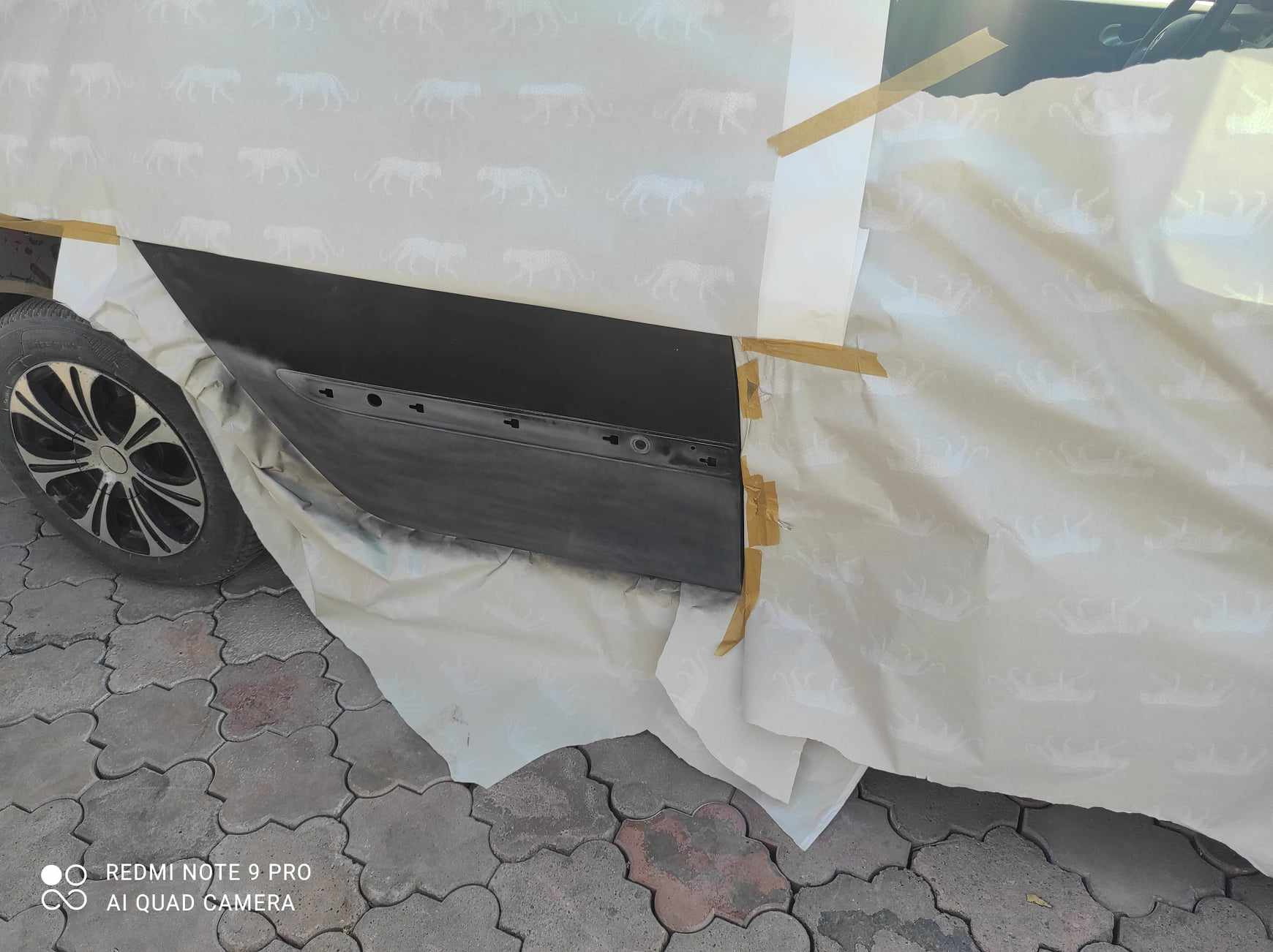 Przygotowanie drzwi samochodu do lakierowania, oklejone papierem ochronnym z motywem gepardów, widoczny fragment opony i kostki brukowej.