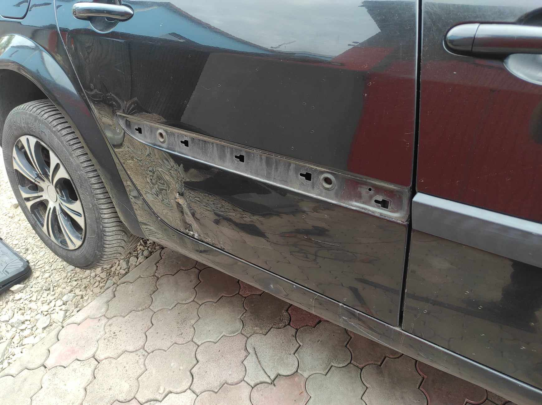 Uszkodzony bok czarnego samochodu z widocznym wgnieceniem i zdemontowaną listwą, przygotowany do naprawy blacharskiej, na kostce brukowej.