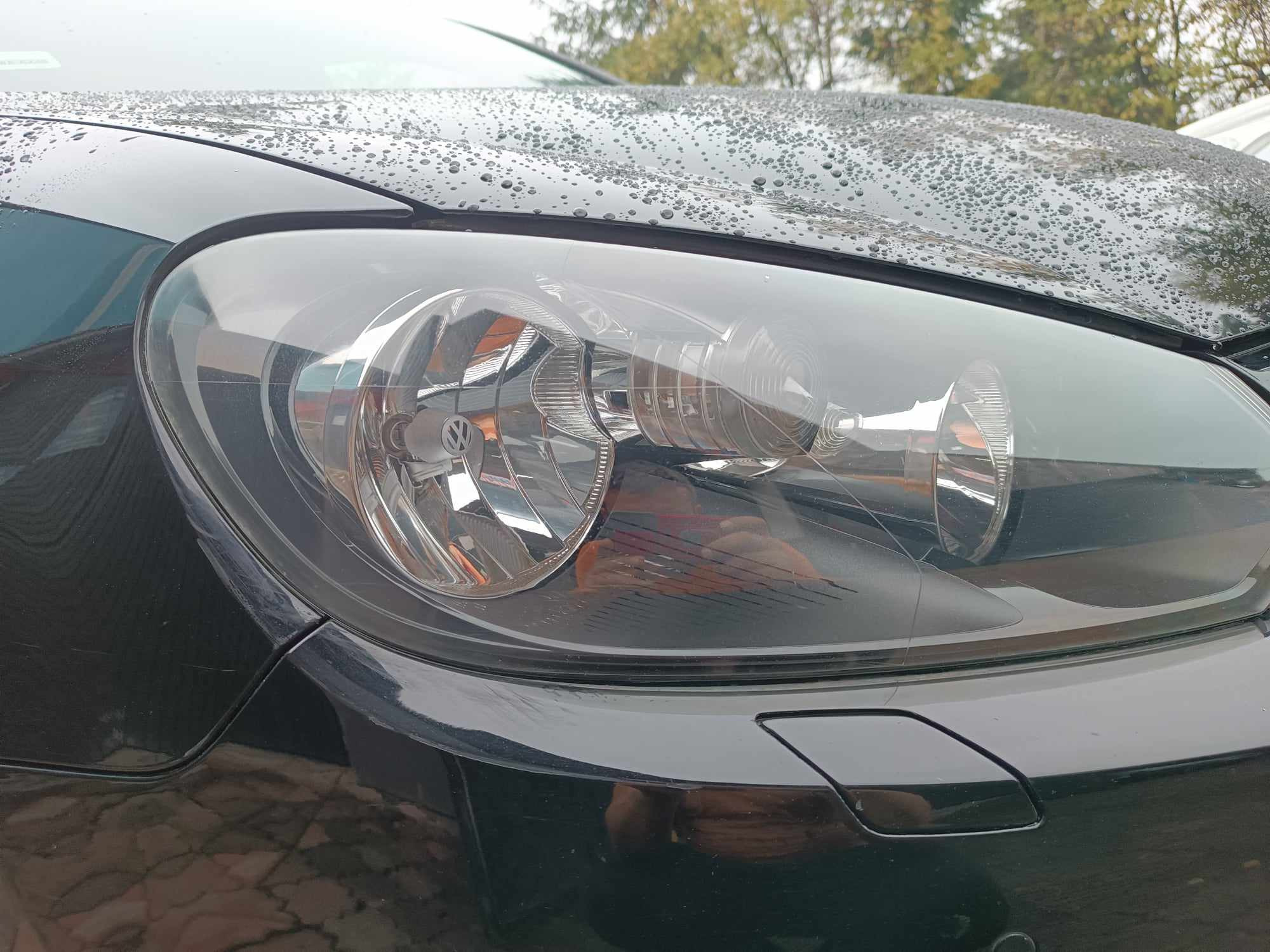 Czarny samochód marki Volkswagen, mokry od deszczu, z widocznym reflektorem i fragmentem karoserii. Na lakierze widoczne liczne krople wody.