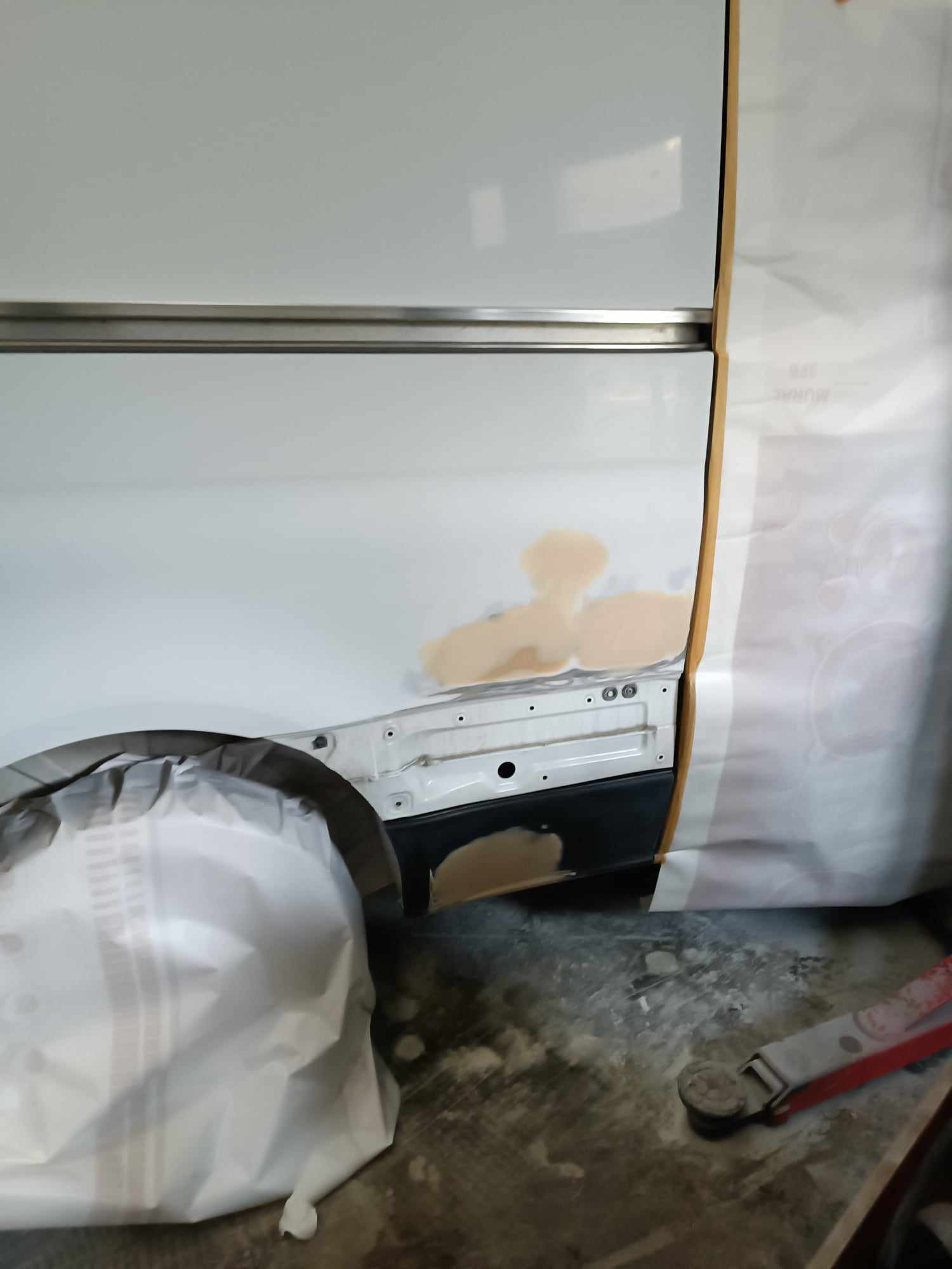 Przygotowanie karoserii samochodu do lakierowania: fragment boku pojazdu z nałożoną szpachlą, zaklejony taśmą i folią ochronną, widoczny podnośnik warsztatowy.