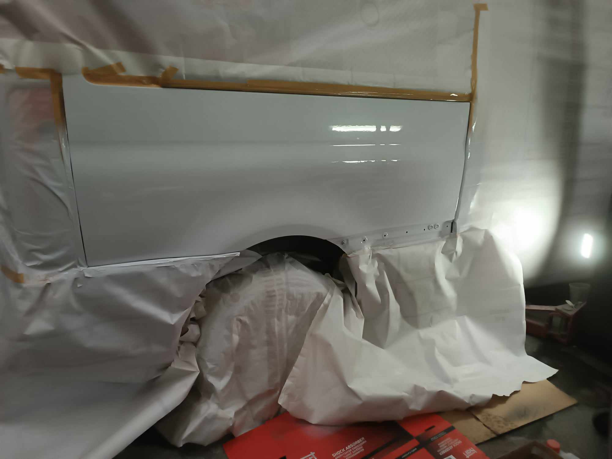 Biały bok samochodu zabezpieczony folią i taśmą malarską przed lakierowaniem, widoczne ślady po naprawach blacharskich.