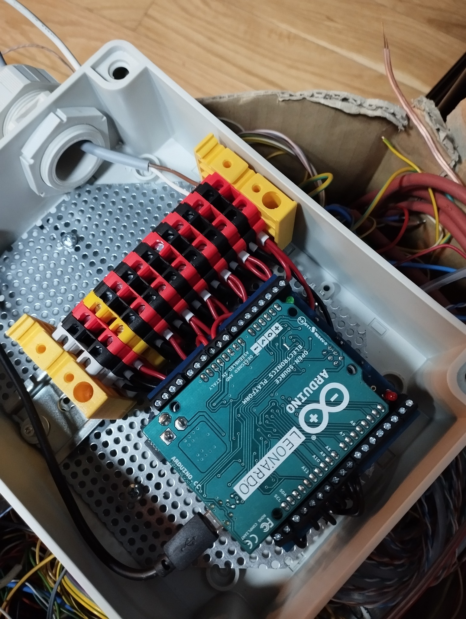 Wnętrze białej skrzynki elektrycznej z perforowanym dnem, zawierającej podłączoną płytkę Arduino Leonardo, listwę zaciskową z przewodami w czarno-czerwonej izolacji i żółte złączki. Widoczne...