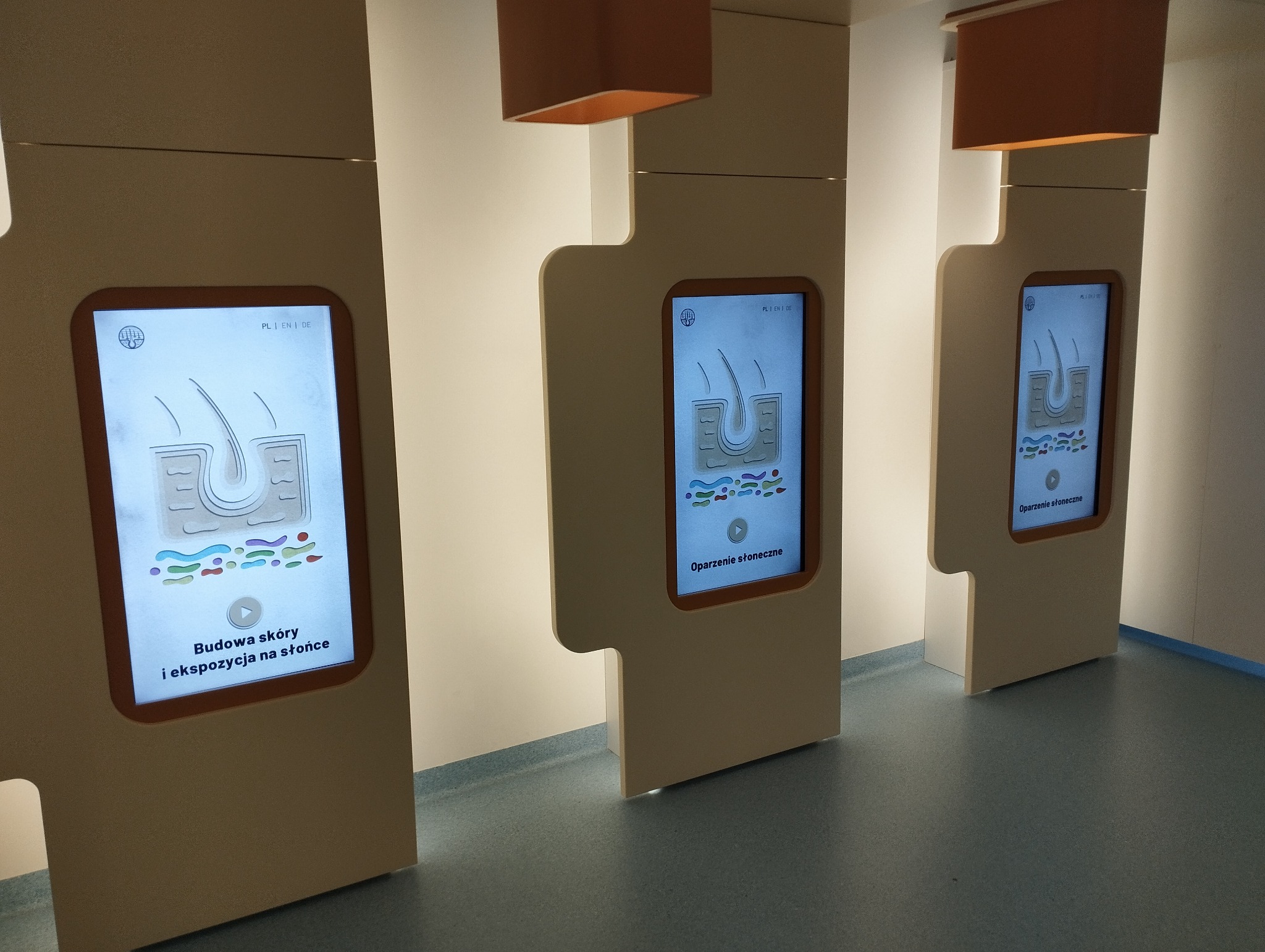 Interaktywne ekrany prezentujące budowę skóry i wpływ ekspozycji na słońce, umieszczone w jasnym pomieszczeniu z minimalistycznym designem.