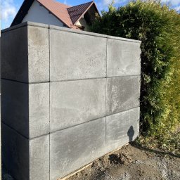 Betonowy ogród - donice, ogrodzenia, kule - z betonu architektonicznego - Tarnów i okolice - Fragment ogrodzenia modułowego z szarego betonu architektonicznego, widoczny z bliska, na tle zielonych krzewów i fragmentu domu z brązowym dachem, cień osoby na ziemi.