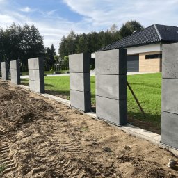 Betonowy ogród - donice, ogrodzenia, kule - z betonu architektonicznego - Tarnów i okolice - Surowe, betonowe słupki ogrodzeniowe w trakcie montażu, widoczne ślady opon na ziemi, nowoczesny dom z ciemnym dachem w tle.