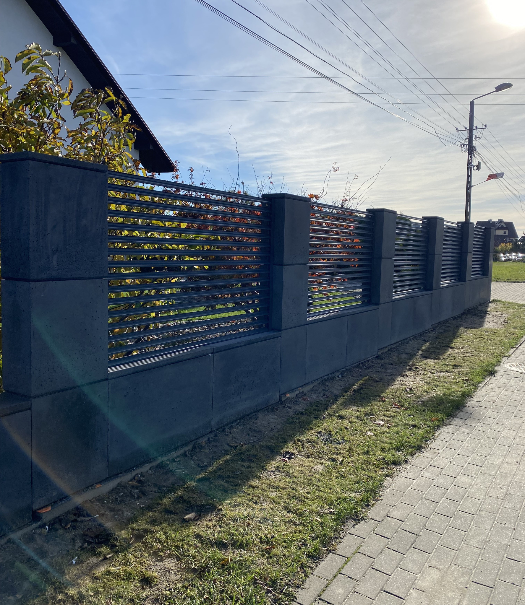 Nowoczesne ogrodzenie panelowe z elementami betonowymi w kolorze grafitowym, widok z poziomu chodnika, jesienne liście w tle.