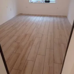 Ułożona podłoga z płytek imitujących drewno w jasnym, pustym pokoju z białymi ścianami i oknem, widoczne krzyżyki dystansowe między płytkami.