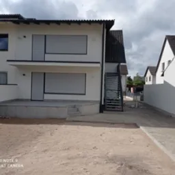 Dwupiętrowy dom z białymi ścianami i szarymi roletami zewnętrznymi, widok z perspektywy niezagospodarowanego terenu przed budynkiem.