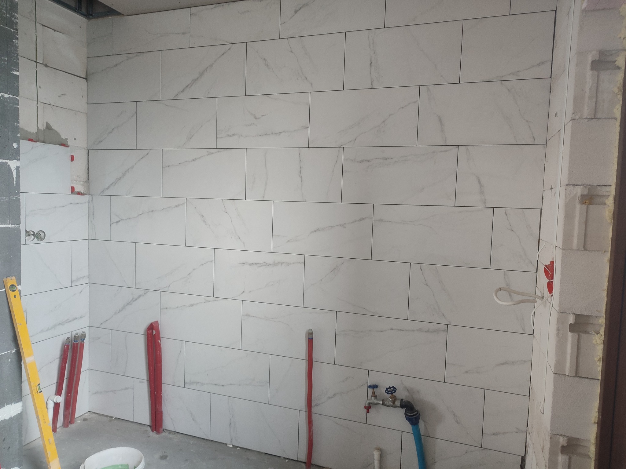 Ściana w trakcie układania płytek imitujących marmur, widoczne rury, poziomica i wiadro z zaprawą.