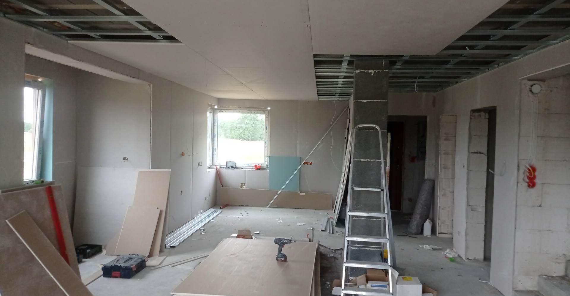 Wnętrze w trakcie montażu płyt karton gips na ścianach i suficie, widoczna metalowa konstrukcja sufitu podwieszanego, drabina i narzędzia.