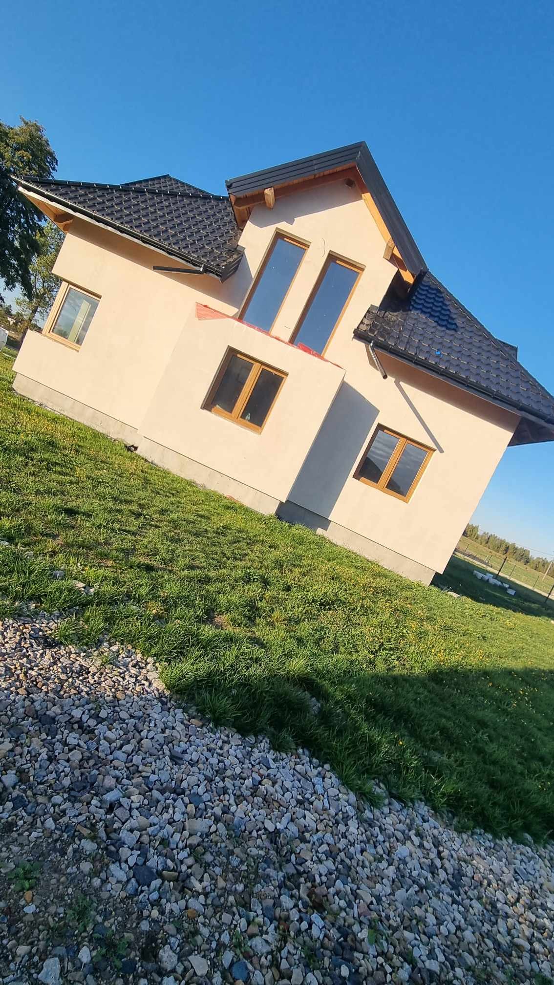 Nowo wybudowany dom jednorodzinny z jasną elewacją i ciemną dachówką, widok z poziomu gruntu na trawnik i kamienie.