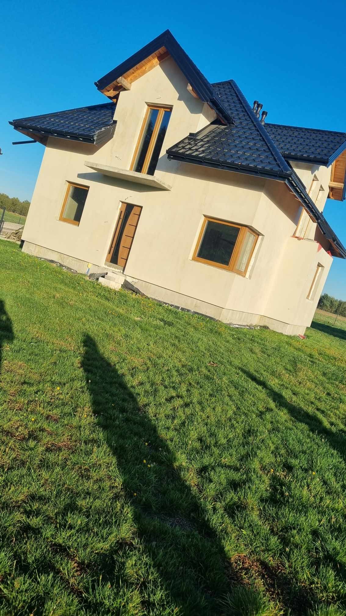 Niedokończony dom jednorodzinny z jasną elewacją, czarnym dachem i drewnianymi oknami na tle zielonego trawnika i błękitnego nieba.