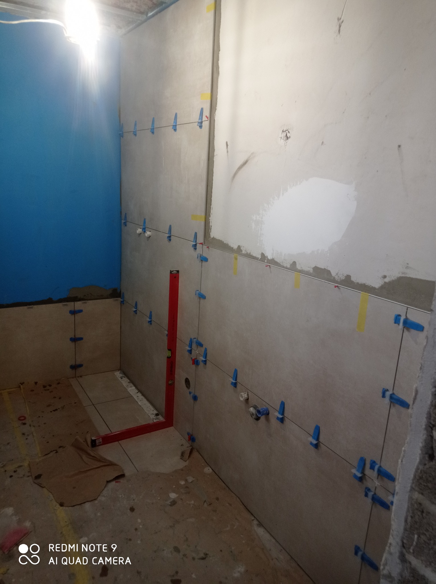 Montaż płytek ściennych w łazience, widoczne niebieskie klipsy poziomujące, poziomica oraz fragment ściany pomalowanej na niebiesko.