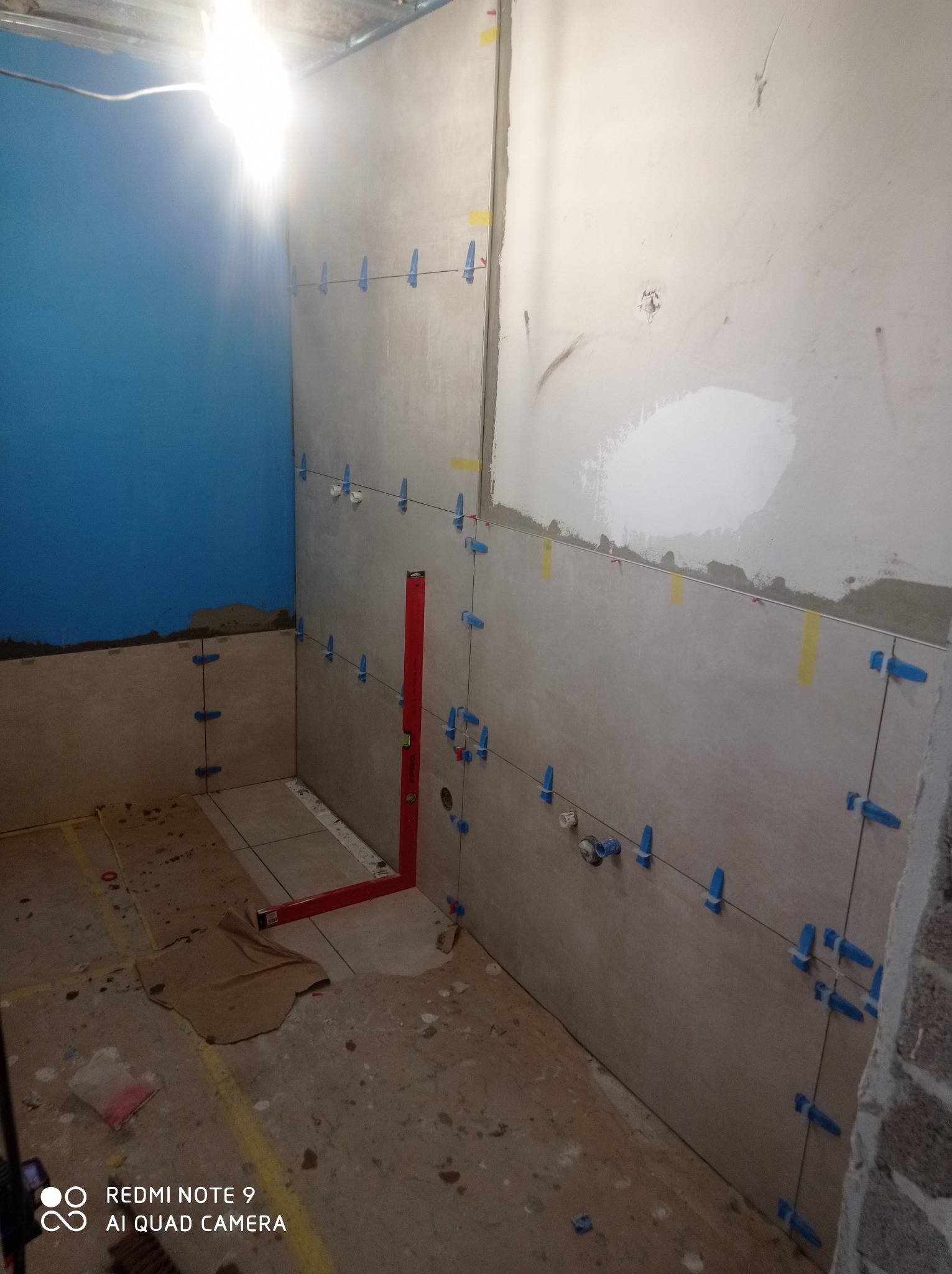 Wnętrze łazienki w trakcie układania płytek ściennych, z użyciem poziomicy i systemu poziomowania z niebieskimi klipsami, na podłodze widoczne zabrudzenia po remoncie.