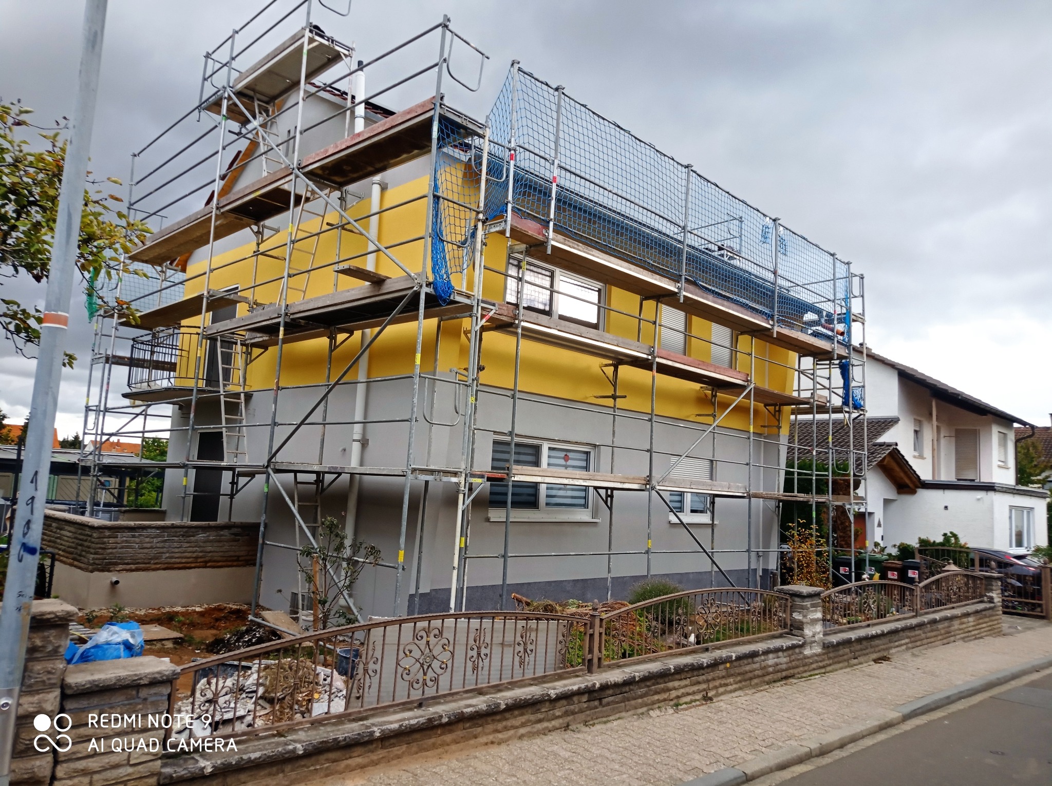 Dom jednorodzinny w trakcie wykonywania elewacji, widoczne rusztowania, siatka zabezpieczająca i fragmenty nowej, żółtej elewacji kontrastujące z szarą, częściowo otynkowaną ścianą.