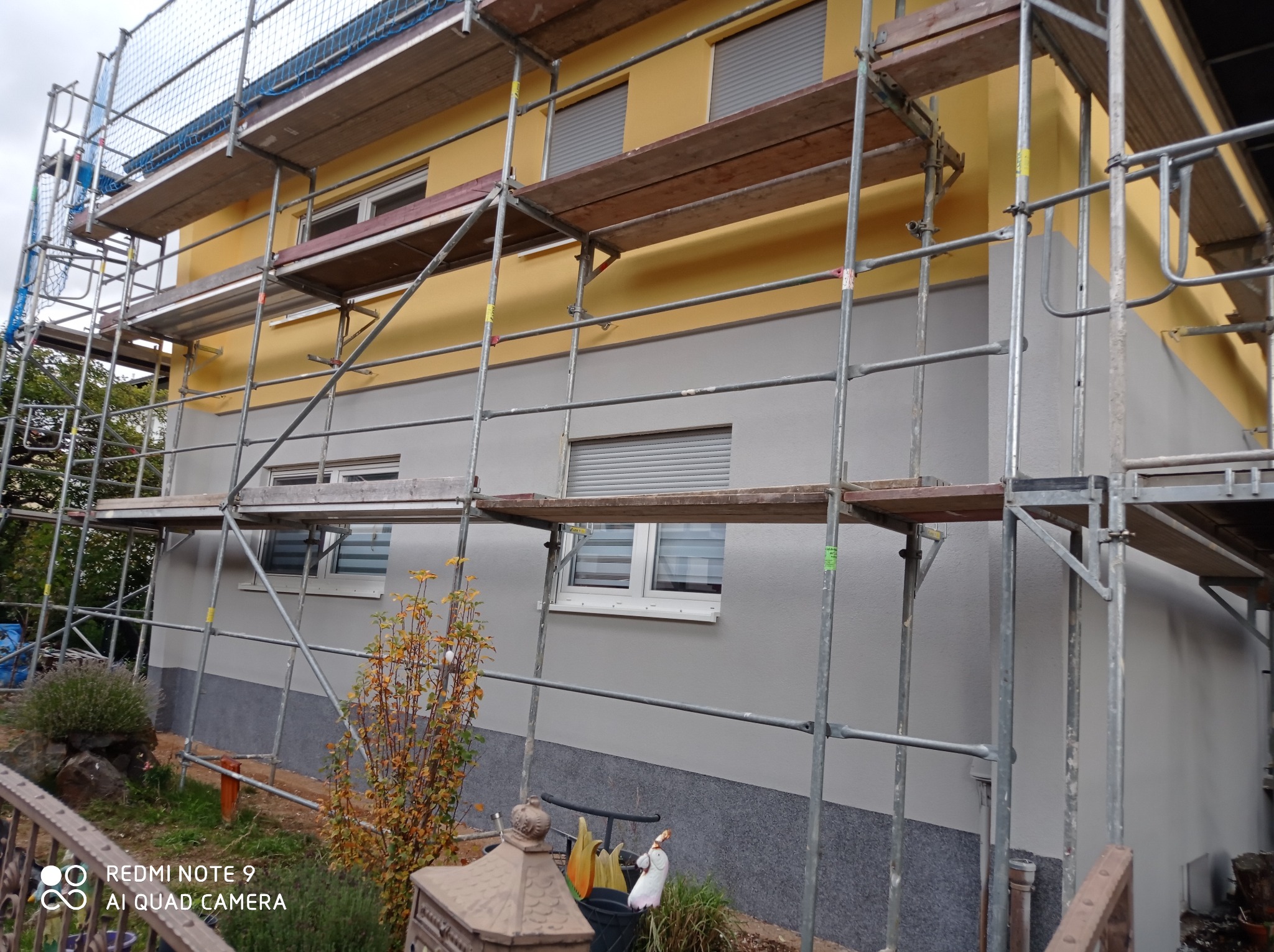 Dwukolorowa elewacja domu jednorodzinnego w trakcie prac wykończeniowych, widoczne rusztowania i częściowo opuszczone rolety okienne.