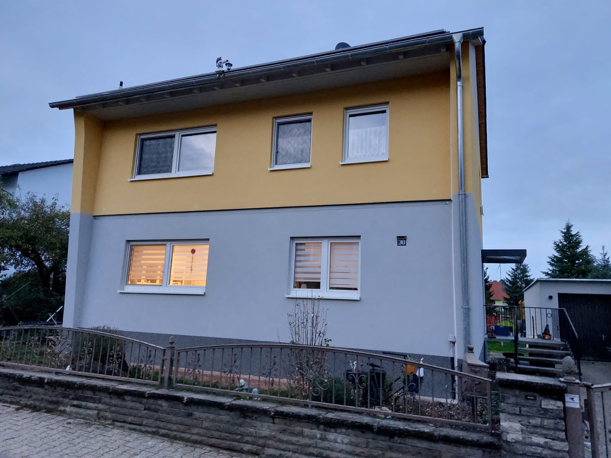 Dwukondygnacyjny dom z żółtą górną częścią i szarą dolną częścią elewacji, widoczny z poziomu ulicy, z numerem 38 na ścianie.