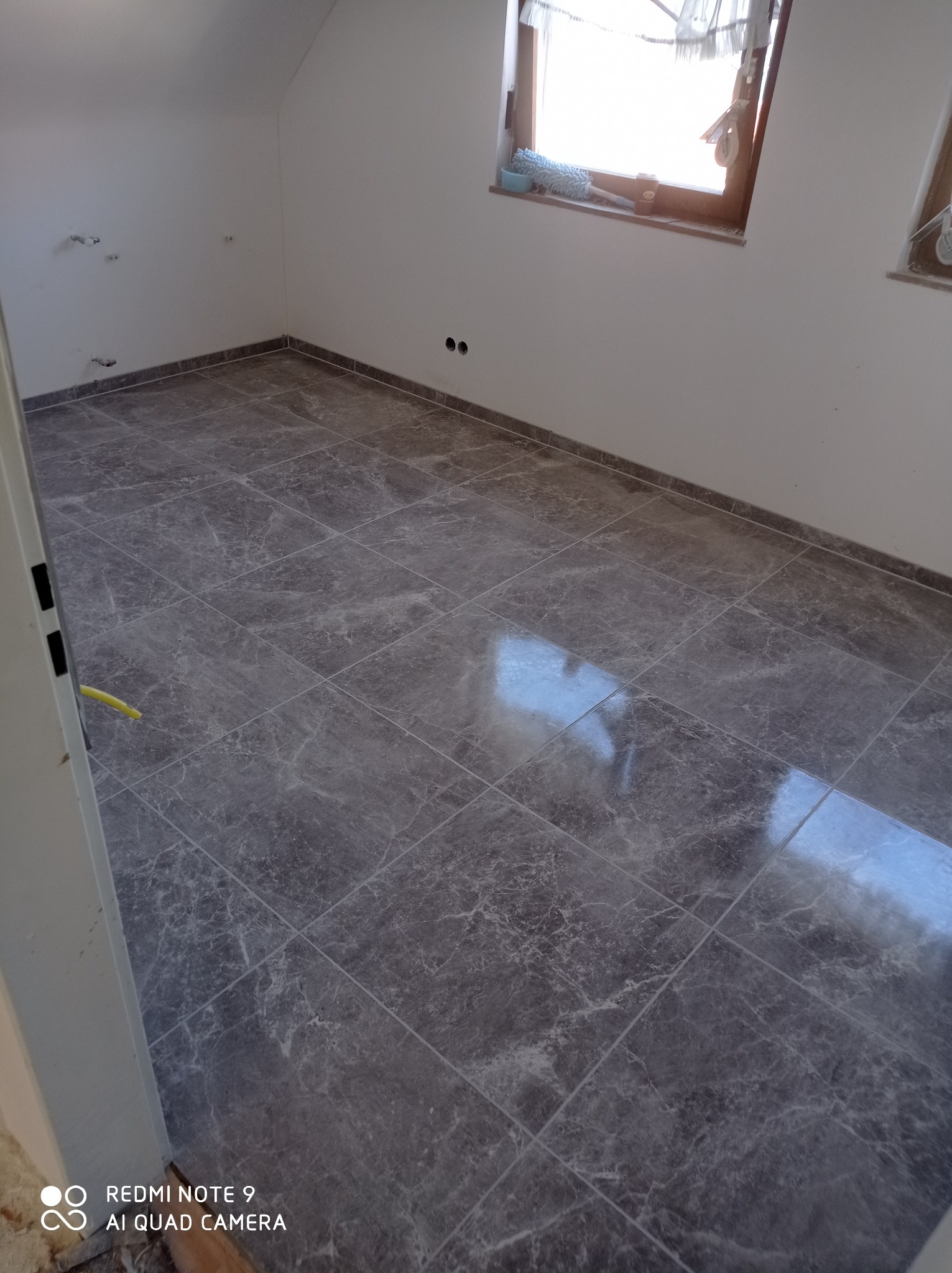 Ułożona szara marmurkowa podłoga z płytek w nowym, pustym pokoju z oknami i żółtym kablem wystającym ze ściany.