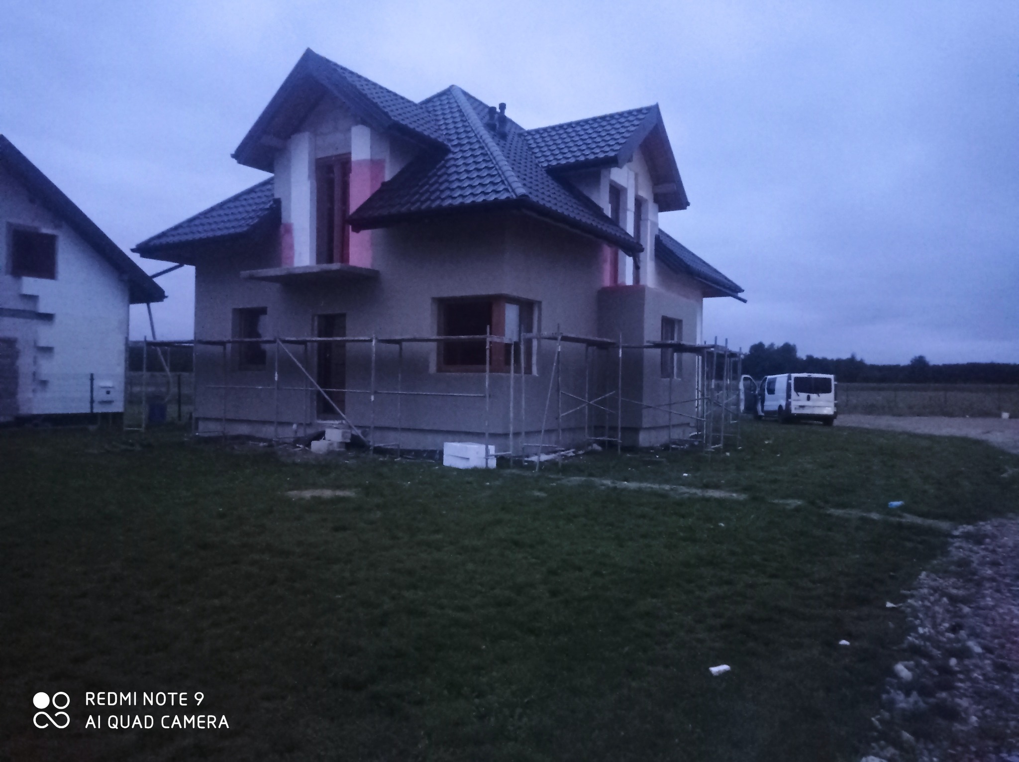 Dom jednorodzinny w trakcie prac elewacyjnych, widoczne rusztowania, częściowo otynkowana fasada, fragmenty ocieplenia, dach pokryty dachówką, na posesji zaparkowany biały bus.