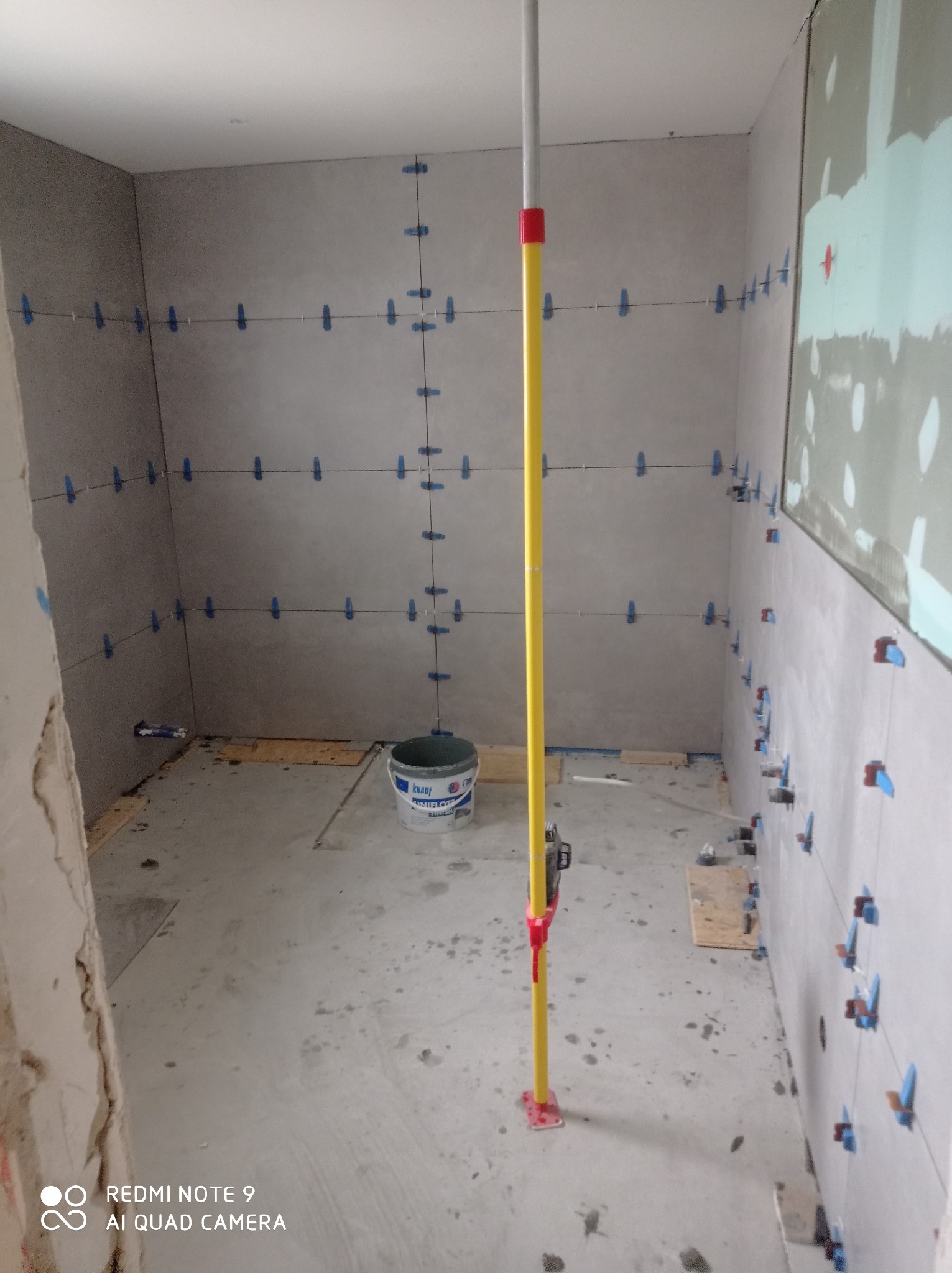 Ściany w trakcie układania płytek ceramicznych w łazience, widoczne krzyżyki dystansowe, poziomica teleskopowa i wiadro z klejem Knauf Uniflot.