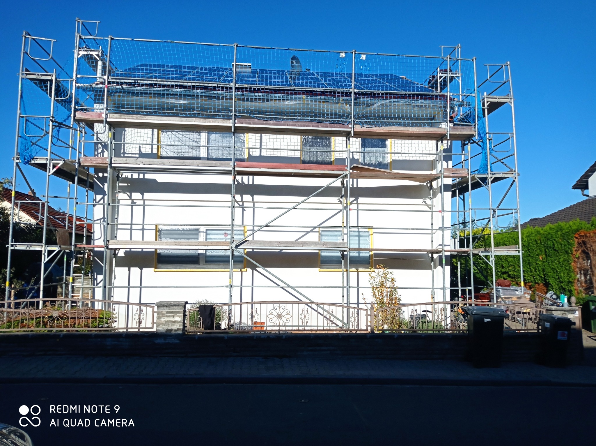 Dwupiętrowy dom w trakcie prac elewacyjnych, otoczony rusztowaniem z niebieską siatką zabezpieczającą, okna zabezpieczone folią.