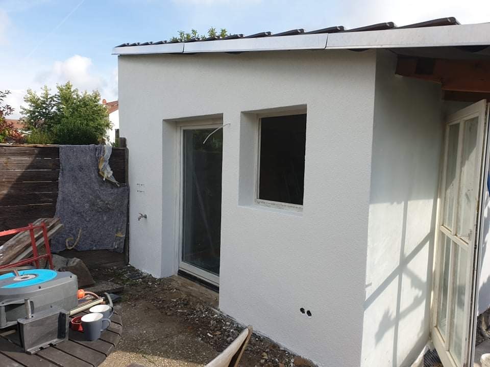 Świeżo otynkowana, biała ściana budynku z dwoma otworami okiennymi i drzwiami, widoczne ślady prac wykończeniowych, narzędzia i materiały budowlane w tle.