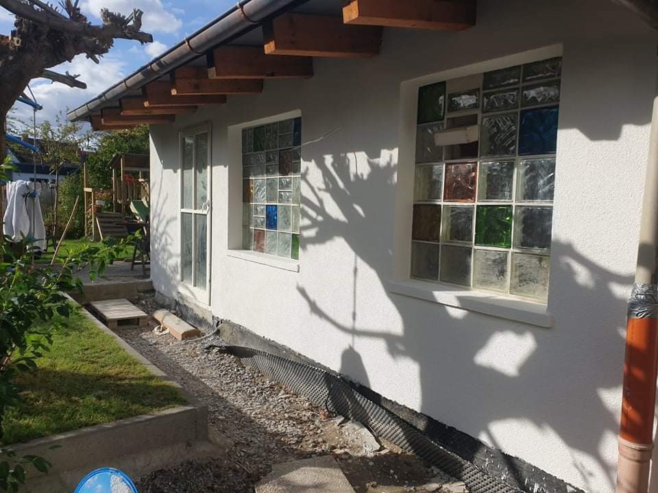 Boczny widok budynku z nową, jasną elewacją, oknami z luksferami w różnych kolorach i zabezpieczeniem fundamentów przed wilgocią.