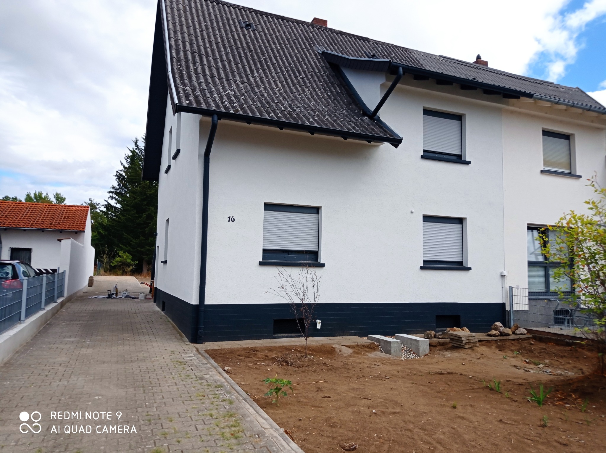 Biały dom dwukondygnacyjny z ciemną podmurówką i szarym dachem, widok z boku na świeżo wyrównany teren przed budynkiem.