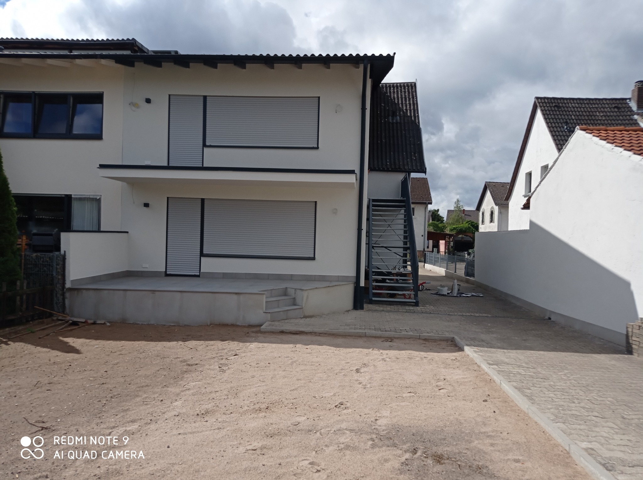 Dwupiętrowy dom z białymi ścianami i szarymi roletami zewnętrznymi, widok z perspektywy niezagospodarowanego terenu przed budynkiem.