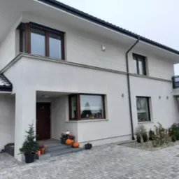 Nowoczesny dom jednorodzinny z jasną, gładką elewacją, brązowymi oknami, szarą dachówką z resztkami śniegu oraz kostką brukową na podjeździe, udekorowany świąteczną choinką i dyniami przy wejściu.