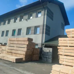 Stosy świeżo przetartego drewna ułożone przed budynkiem z szarymi paskami na fasadzie i błękitnym niebem w tle.