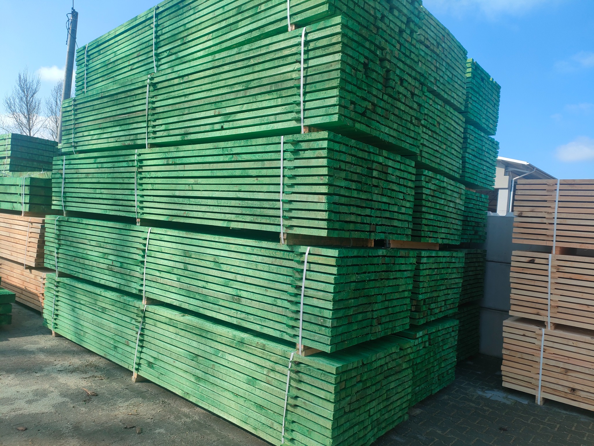 Stosy świeżo impregnowanej na zielono tarcicy budowlanej, zabezpieczone białymi taśmami, ułożone na placu składowym.