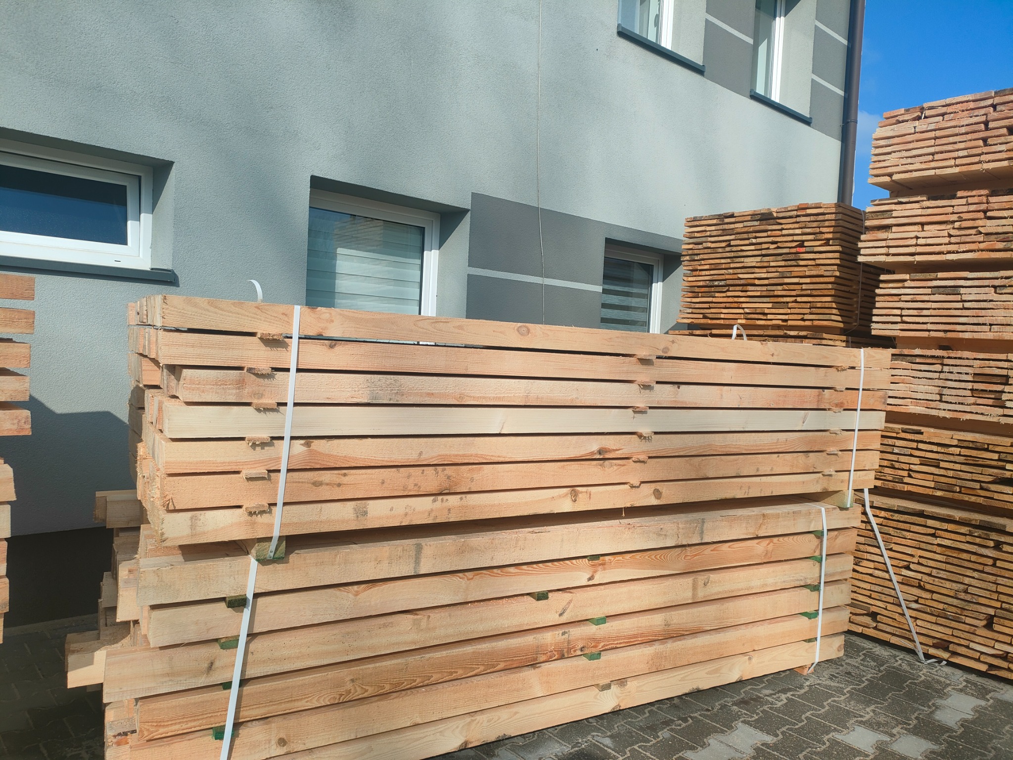 Stosy świeżo ściętej tarcicy budowlanej, zabezpieczone białymi taśmami, ułożone na brukowanym placu przed budynkiem w odcieniach szarości.