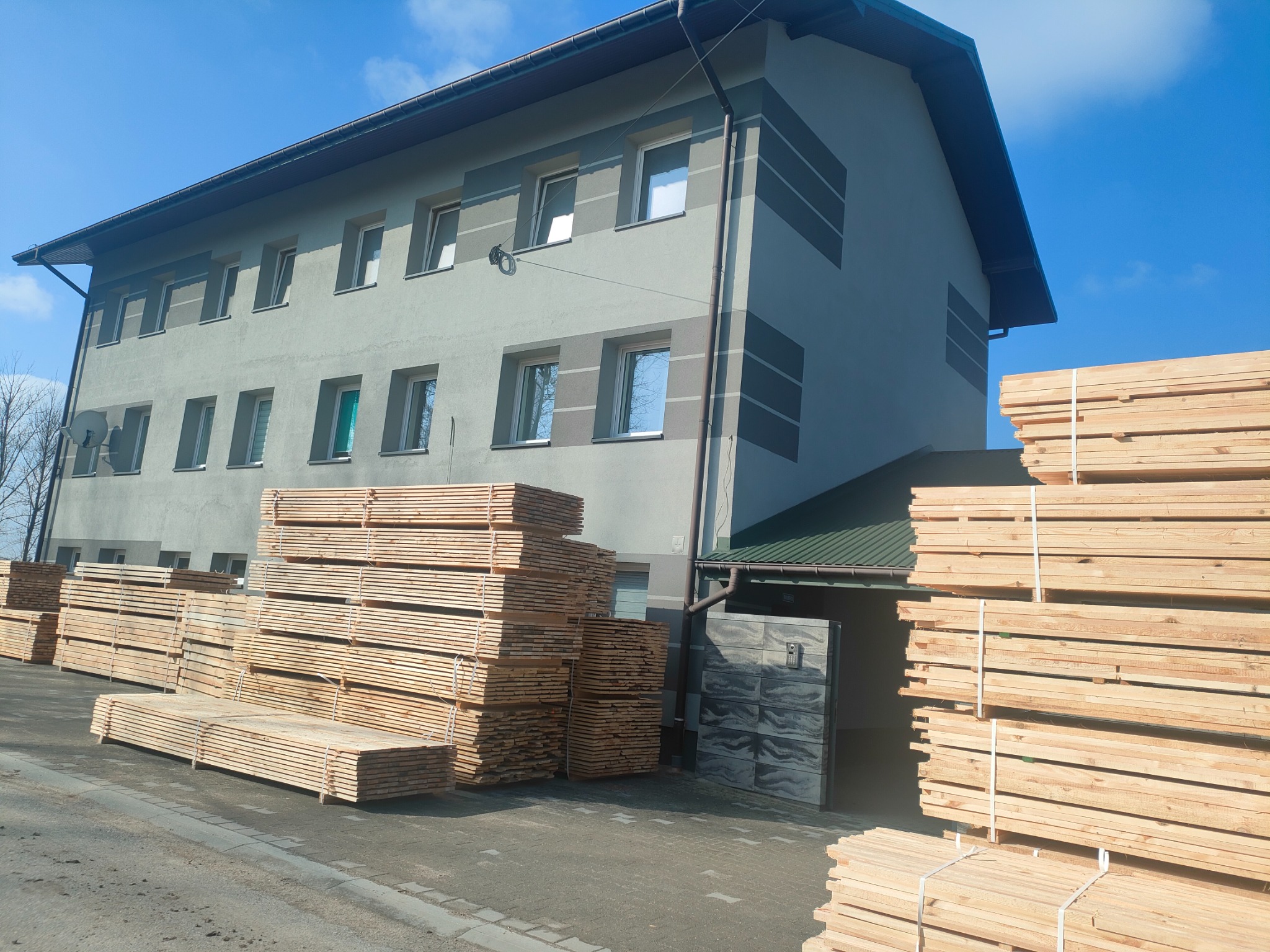 Stosy świeżo przetartego drewna ułożone przed budynkiem z szarymi paskami na fasadzie i błękitnym niebem w tle.