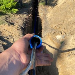 Wiktor Frajer - Ręka trzymająca kabel elektryczny w niebieskiej rurze, widoczny wykop w ziemi z niebieską rurą w środku.