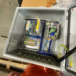 Wiktor Frajer - Otwarta szara szafa sterownicza z widocznymi elementami elektrycznymi, w tym modułami, listwami zaciskowymi i okablowaniem w kolorach niebieskim, żółtym i zielonym, z wentylatorem po lewej stronie.