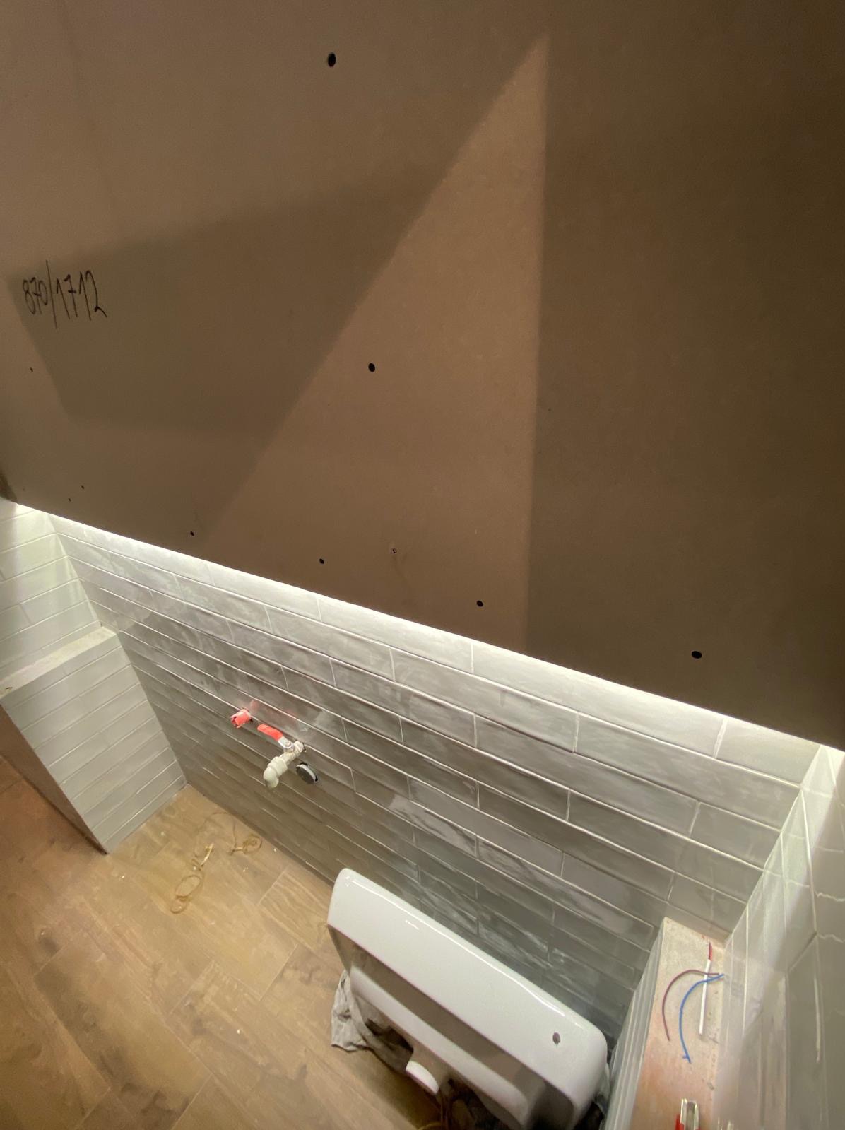 Niedokończona łazienka z szarą glazurą ułożoną poziomo, widoczne wystające rury, podwieszany sufit z oświetleniem LED i odręczny napis '870/1712' na płycie.