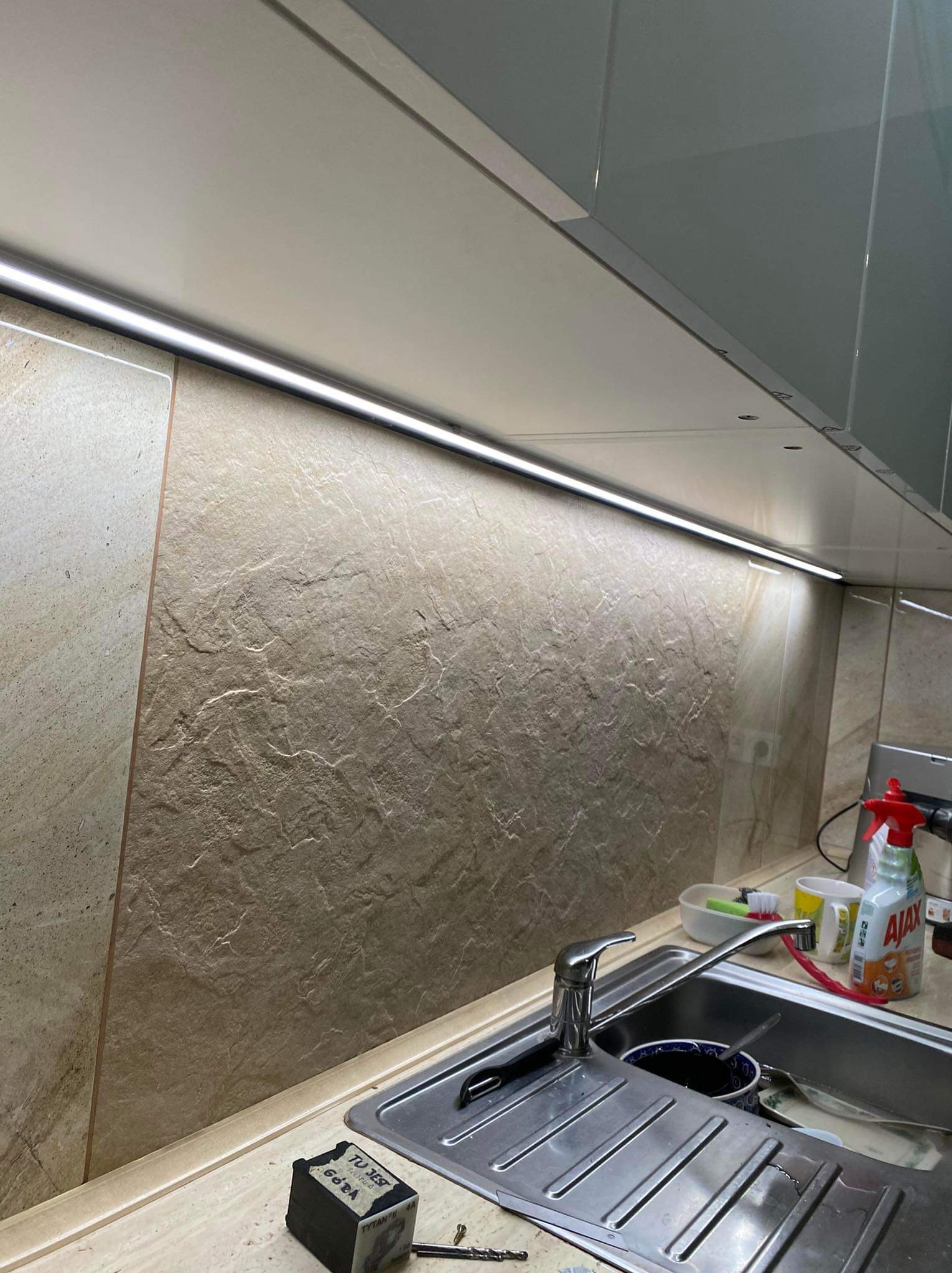 Kuchnia z oświetleniem LED podszafkowym, zlewozmywak ze stali nierdzewnej i dekoracyjna ściana z kamienia imitującego. Widoczny tester napięcia.