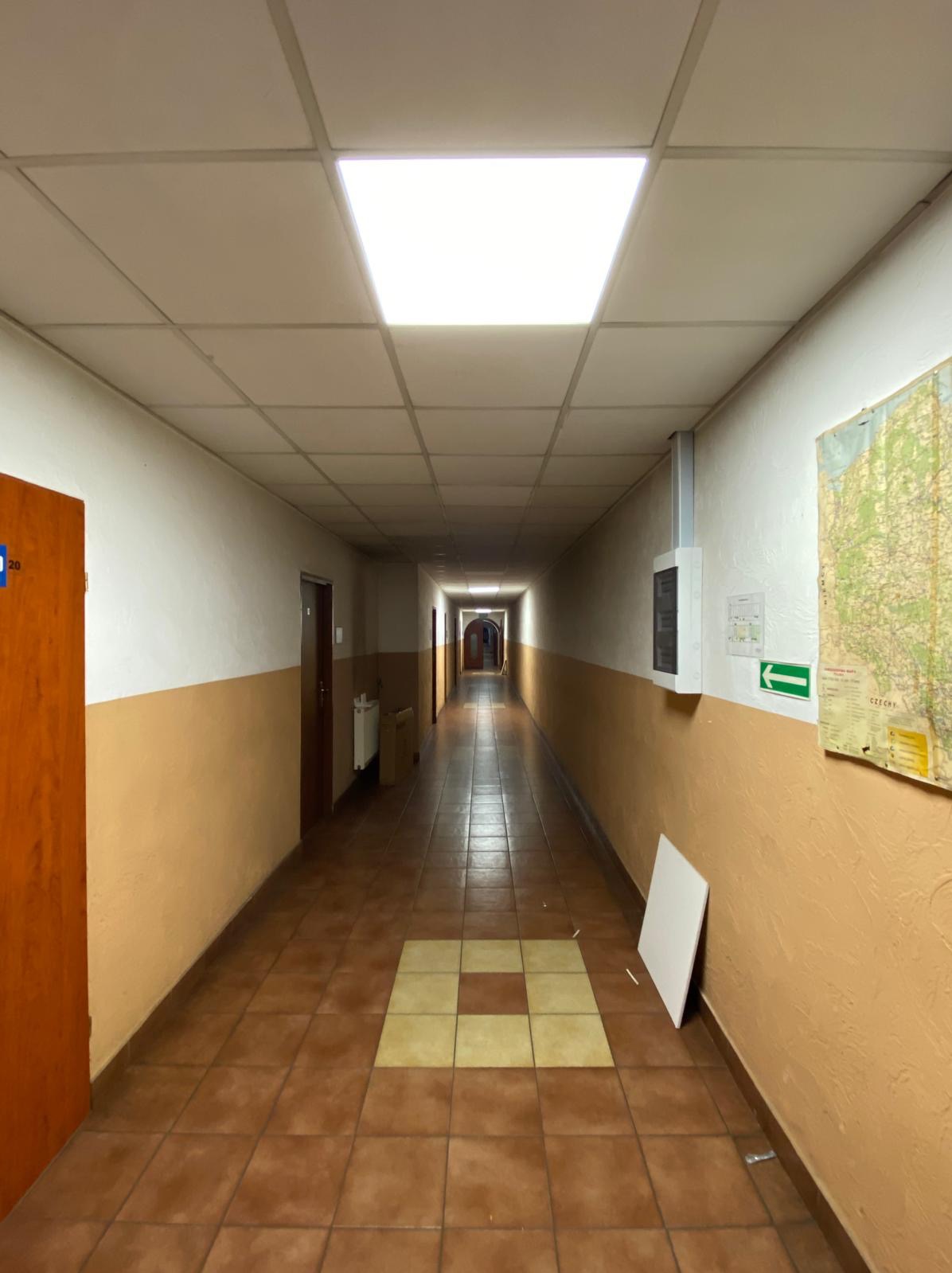 Długi korytarz z brązowymi płytkami podłogowymi i białymi sufitowymi panelami oświetleniowymi, na ścianie tablica rozdzielcza i mapa Europy Środkowej.