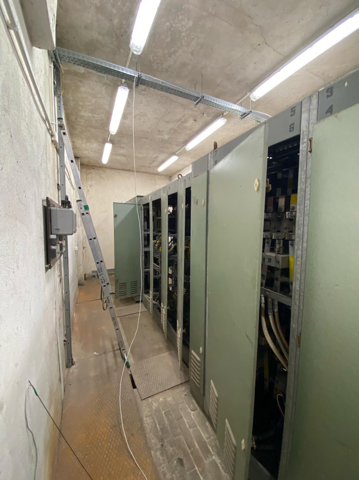 Wnętrze pomieszczenia z rzędem szaf elektrycznych z otwartymi drzwiami, drabina oparta o ścianę, widoczne kable i oświetlenie sufitowe.