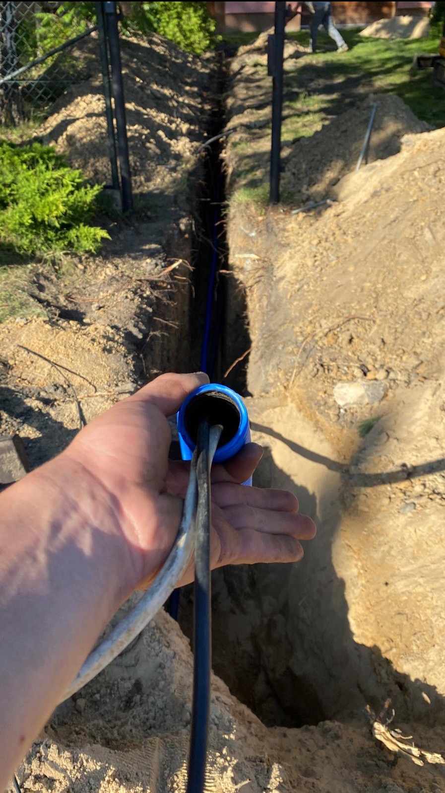 Ręka trzymająca kabel elektryczny w niebieskiej rurze, widoczny wykop w ziemi z niebieską rurą w środku.