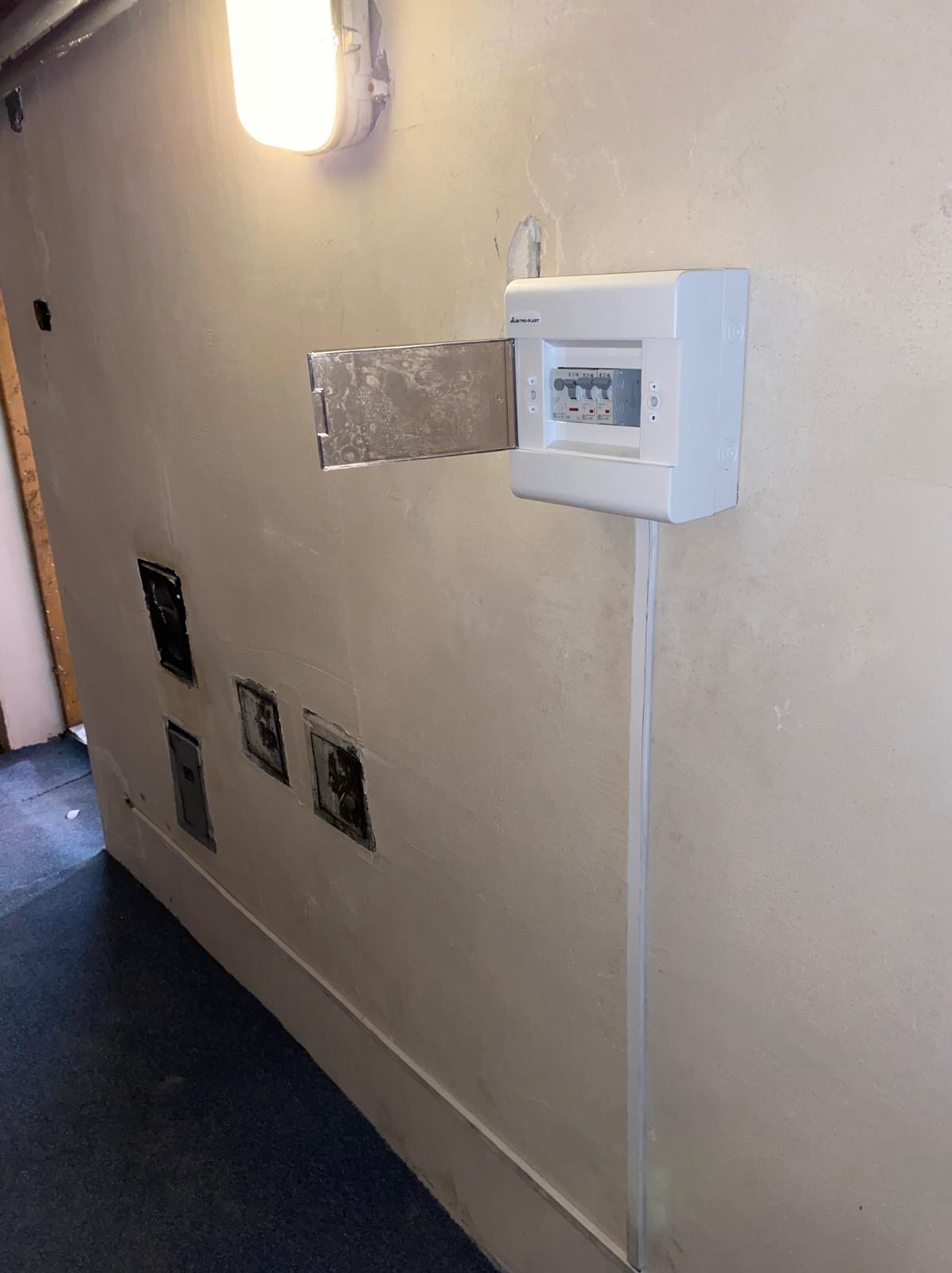 Otwarta skrzynka bezpiecznikowa na ścianie z widocznymi śladami po starych gniazdkach elektrycznych, oświetlona lampą przemysłową.