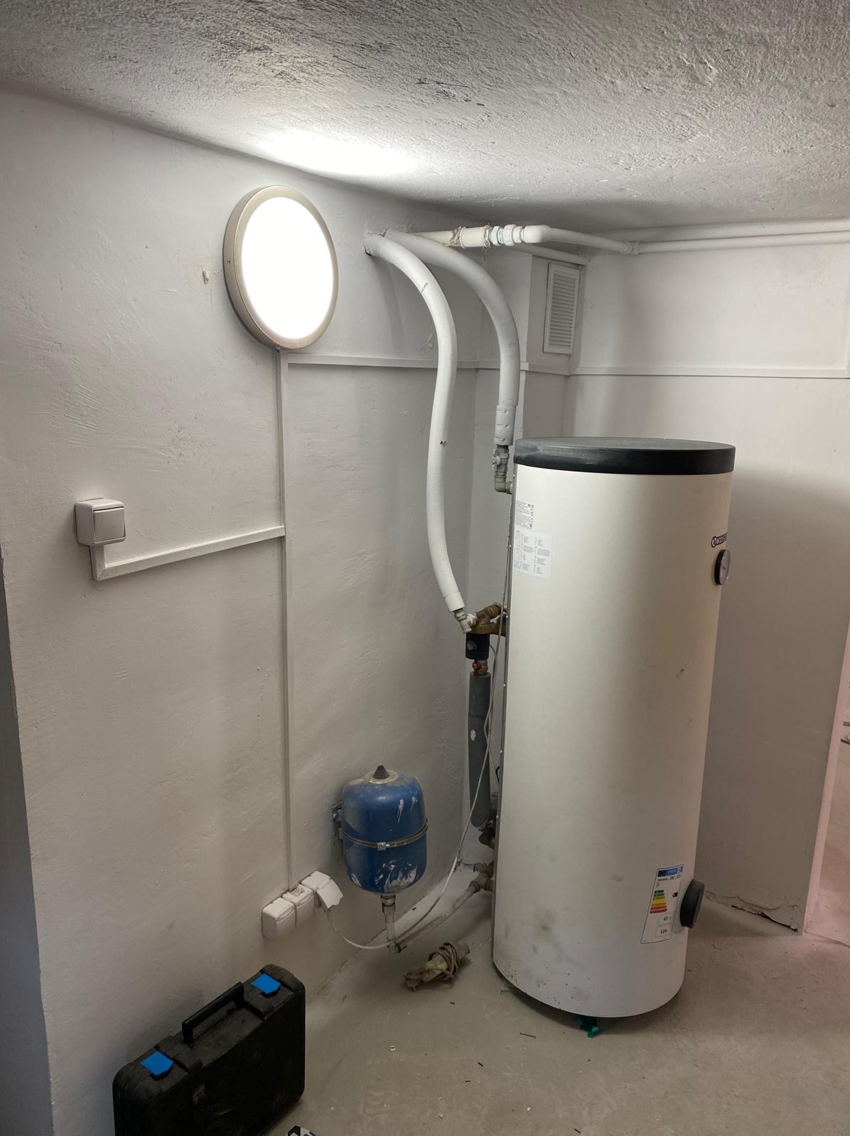 Bojler elektryczny z podłączonym osprzętem w pomieszczeniu technicznym, widoczne rury, zawory, zbiornik wyrównawczy i skrzynka narzędziowa na podłodze.