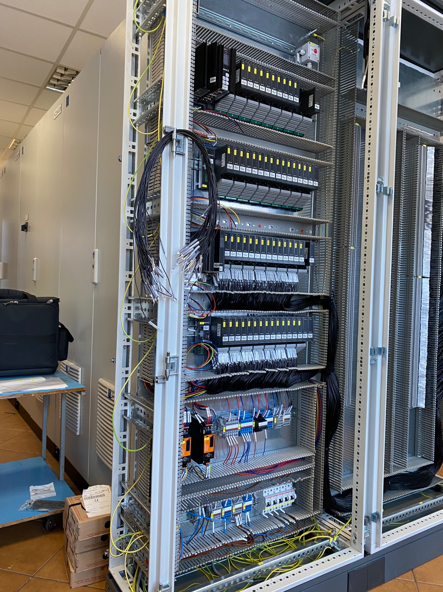 Rozbudowana szafa sterownicza z licznymi modułami, kablami i złączkami, widoczna w pomieszczeniu technicznym, z torbą narzędziową i kartonami w tle.