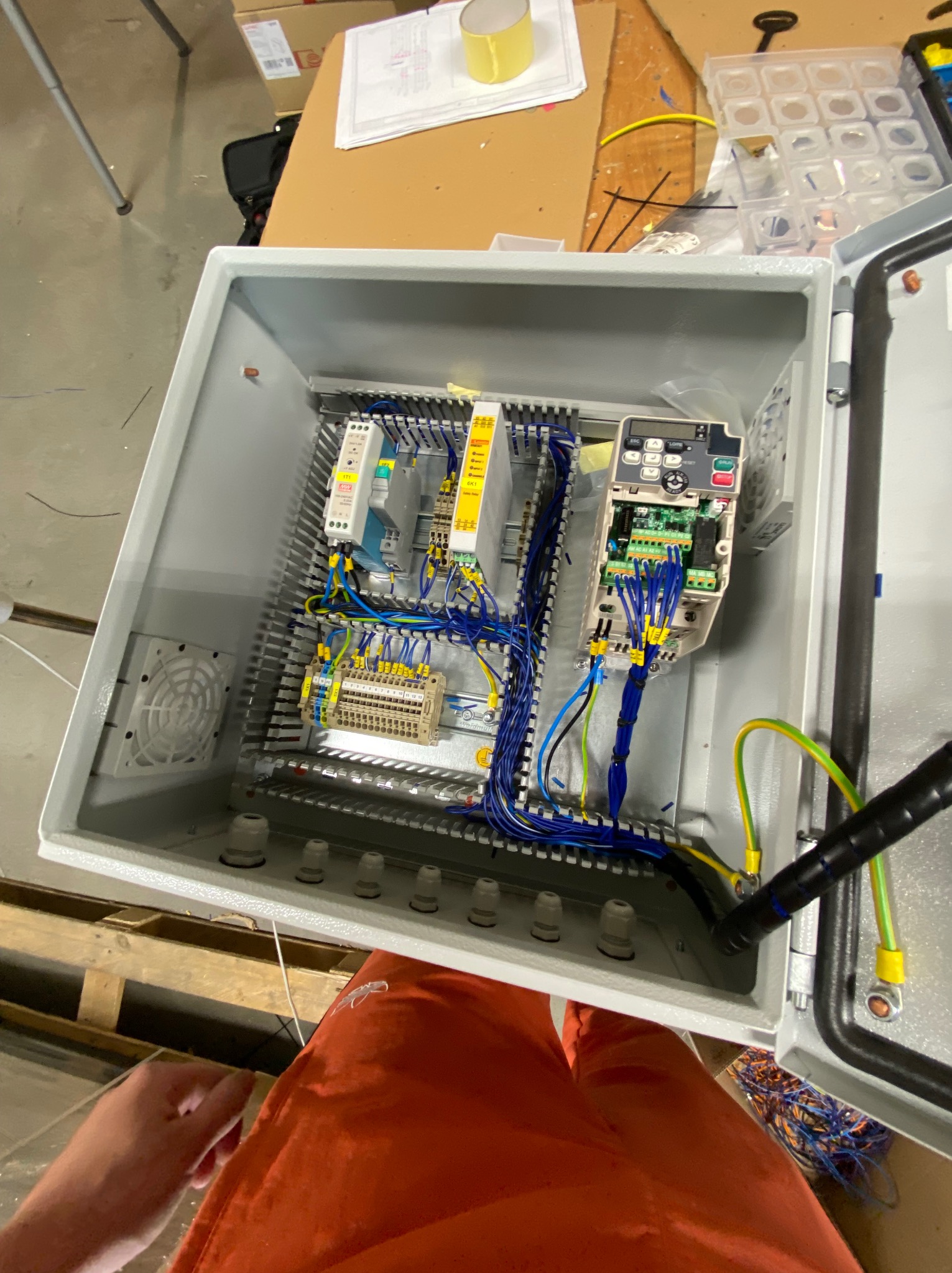 Otwarta szara szafa sterownicza z widocznymi elementami elektrycznymi, w tym modułami, listwami zaciskowymi i okablowaniem w kolorach niebieskim, żółtym i zielonym, z wentylatorem po lewej stronie.