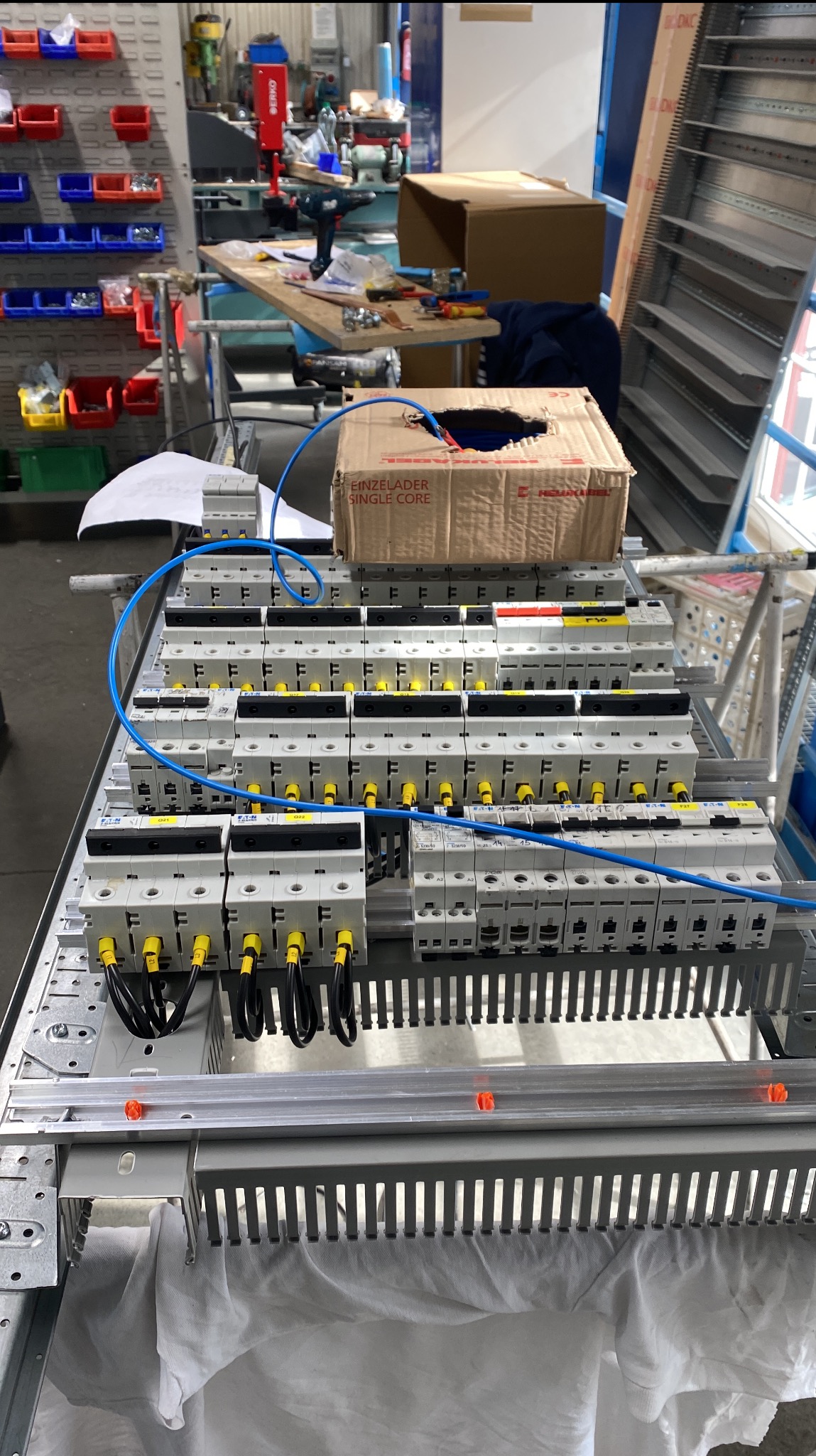 Rozdzielnica elektryczna w trakcie montażu, widoczne zabezpieczenia modułowe, przewody w peszlach i skrzynka kartonowa na górze.