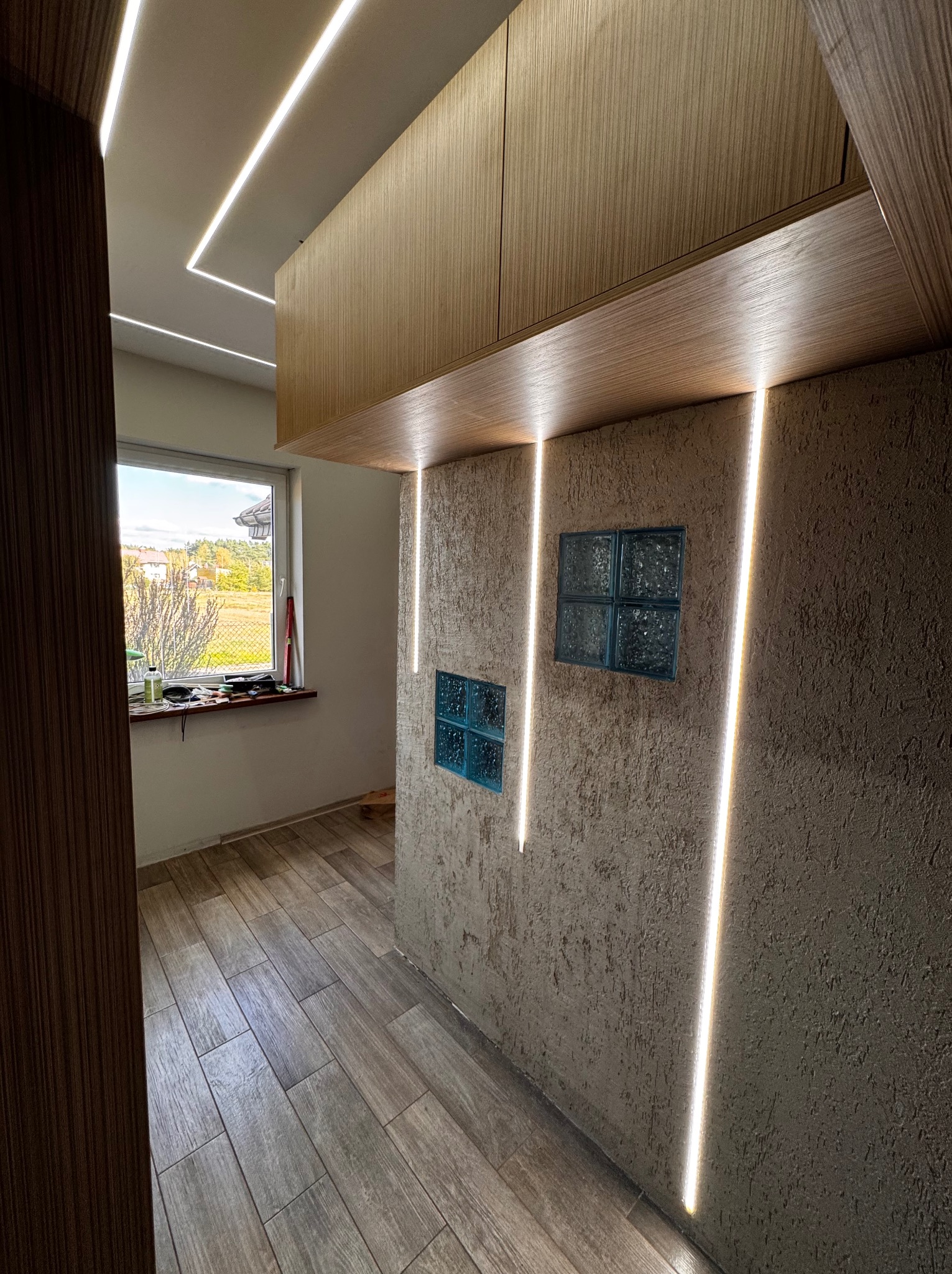 Wnętrze korytarza z drewnianą zabudową, oświetleniem LED w pionowych pasach na ścianie o fakturze baranka oraz szklanymi luksferami, widok na okno z narzędziami na parapecie i krajobraz za nim.
